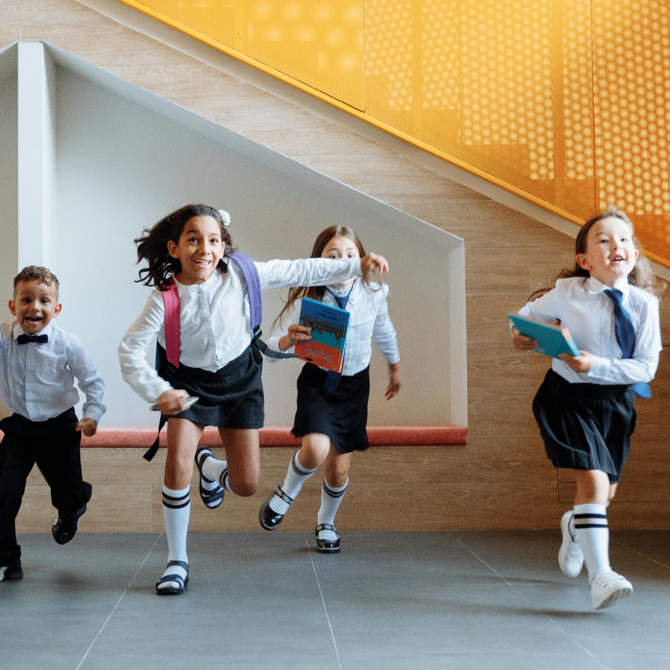 Four children in school uniforms joyfully running up towards camera, holding books.